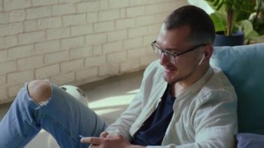 Gözlüklü neşeli bir adam telefonda konuşuyor. Kablosuz kulaklıkları açık apartmanda yerde oturuyor.
