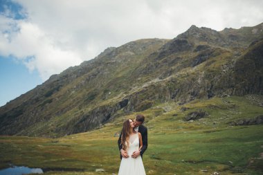 Damat portresi, geline arkadan sarılıp öpüyor. Dağların arkasında poz veriyor. Şık bir portre. Mutlu aile birlikte.