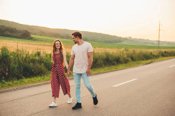 Pareja sonriente caminando por un camino de asfalto cogida de la mano, chica mirando a su amante. Día de la familia. Relación de actividad. Ocio, viajes, turismo. Familia feliz — Foto de Stock