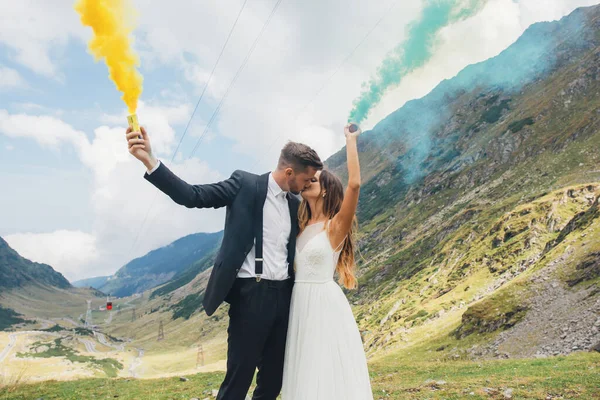 Retrato de um recém-casados com a fumaça de cor amarela e azul nas mãos posando enquanto se beijam na montanha. Natureza paisagem. Dia do casamento. — Fotografia de Stock