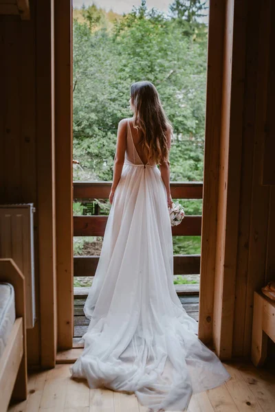 Visão traseira de uma noiva de cabelos castanhos vestida com um longo vestido branco admirando a paisagem da montanha a partir da varanda de uma casa de campo. Bonito para design de conceito — Fotografia de Stock