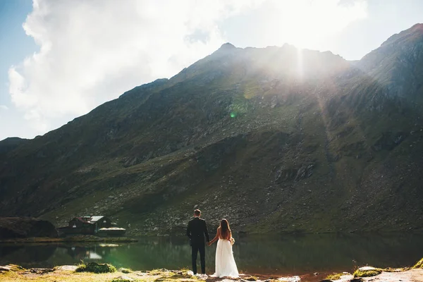 Vista trasera de una pareja de jóvenes casados tomados de la mano y mirando el lago y el hermoso paisaje de montaña que tienen en su boda. Paisaje de montaña —  Fotos de Stock