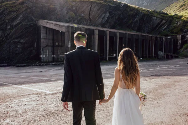 Visão traseira de um casal recém-casado de mãos dadas, noivo em um terno e mulher em um vestido branco longo ir para o estacionamento localizado um penhasco da montanha para levar seu carro e — Fotografia de Stock