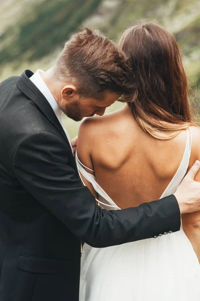 Ritratto ravvicinato di uno sposo che bacia la spalla della sposa in piedi con la schiena davanti alla telecamera indossando un vestito con la schiena scoperta. — Foto Stock