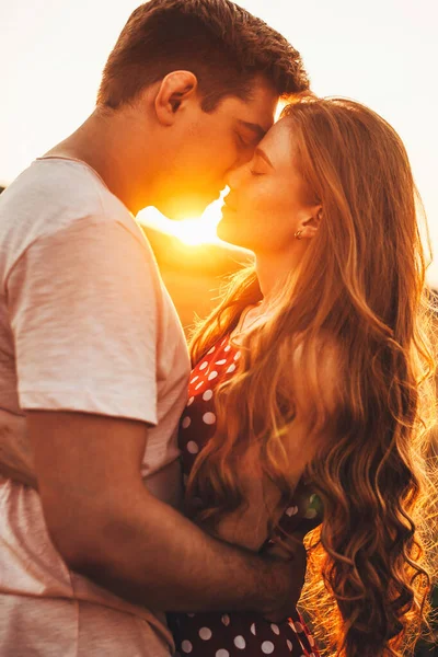 Vista lateral de un par de recién casados de pie y abrazándose ante un beso contra el fondo de la puesta de sol en el campo de trigo. Retrato para el diseño de estilo de vida. —  Fotos de Stock