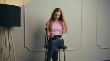 Kızıl saçlı kız, sessiz bir odada, bir sandalyede tek başına oturmuş deftere yazı yazıyor. Güzel bir kadın. Zarif, modern tarz. Beyaz Duvar.