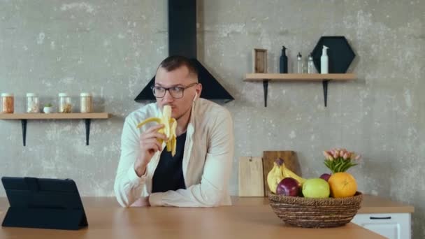 Vit man som står i köket och äter banan medan han pratar med lagkamrater under onlinemötet använder surfplatta. Nätkommunikation — Stockvideo