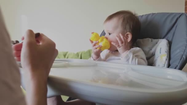 Bebé jugando con un juguete de goma amarillo durante el desayuno, madre alimentando al bebé con cuchara. Cuidado familiar. Cuidado del bebé. — Vídeo de stock