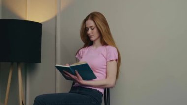 Uzun kızıl saçlı bir kadının portresi, beyaz duvarın üzerinde bir sandalyede oturmuş, bir günlüğe yazı yazıyor. Beyaz duvar. Güzel kız..