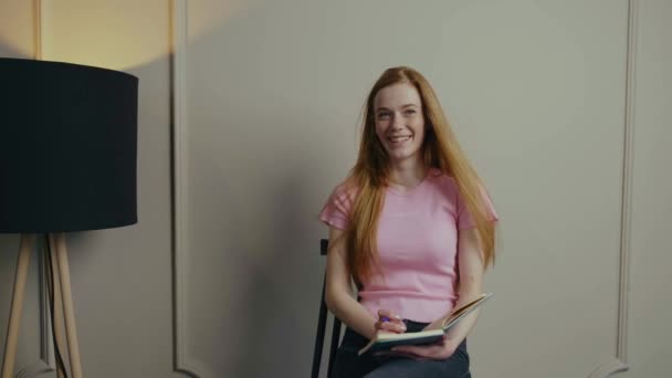 Studentessa dai capelli rossi con copybook sorridente alla macchina fotografica facendo compito di prova mentre posa seduta sulla sedia contro parete bianca. — Video Stock