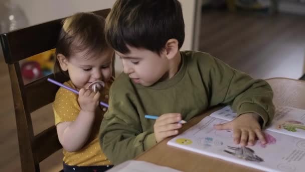 Broder leker med babysister samtidigt som han njuter av fritiden tillsammans hemma och ritar med färgade pennor när han sitter vid bordet. Utbildning för barn. Lyckligt ansikte — Stockvideo