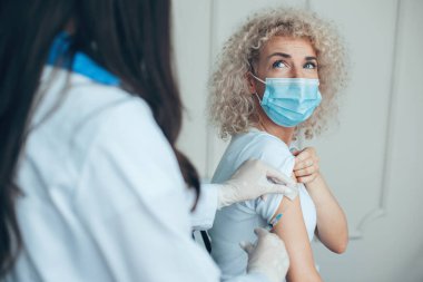 Tıbbi maskeli beyaz kadın modern klinikte aşı enjeksiyonu yaptırıyor. Korkmuş görünüyorsun. Coronavirus güvenliği. Coronavirus aşısı