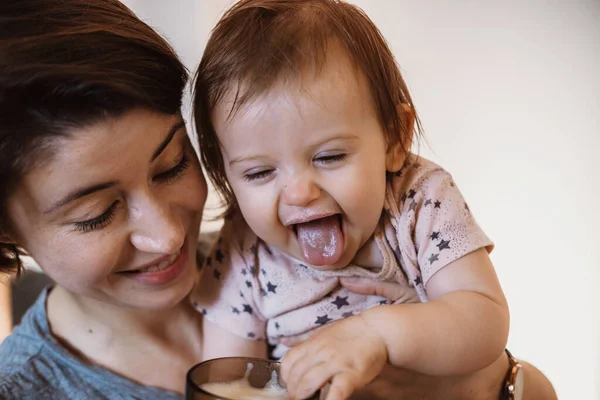 Mamman håller barnet i sina armar och håller ut ett glas mjölk att dricka, barnet sticker ut tungan. Familjeomsorg. Barnomsorg. — Stockfoto