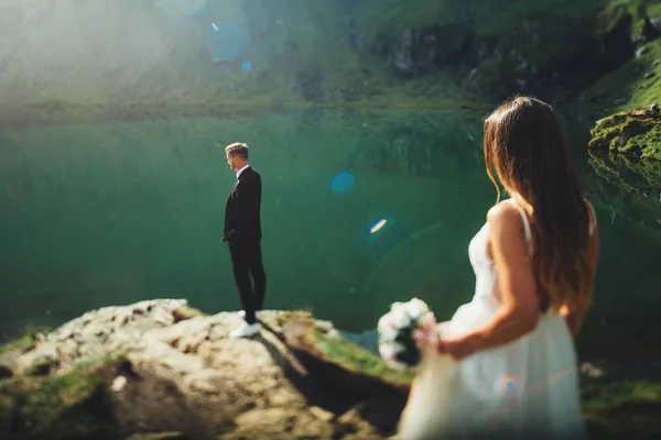 Novia celebración de un hermoso ramo de flores blancas de la boda va al novio que la espera de pie cerca del lago y piedras edificios. Día de la boda. —  Fotos de Stock