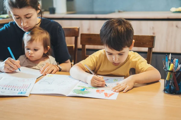 Niño sentado en la mesa y practicando la escritura, madre sosteniendo a la niña escribiendo enfocada en una sábana. Educación infantil. Estudiar, hacer los deberes. —  Fotos de Stock