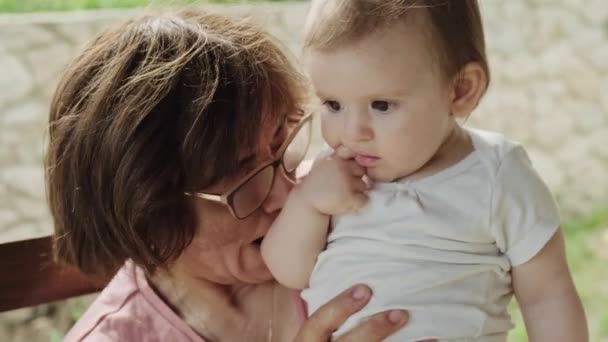 Nonna seduta fuori sulla panchina nel cortile e giocare con la nipote baciarla mentre la tiene in braccio. Nonna tenendo il bambino all'aperto, grande — Video Stock