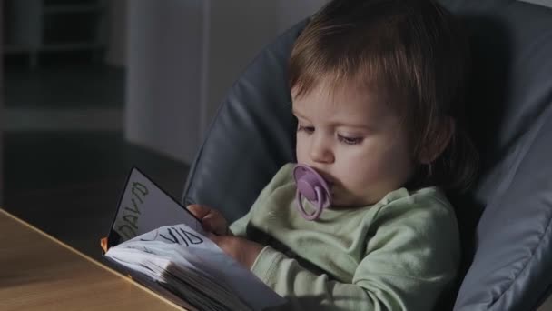 Curiosa bambina seduta su un seggiolone a sfogliare le pagine del diario dei suoi fratelli tenendo in bocca un ciuccio. Libro per bambini. — Video Stock