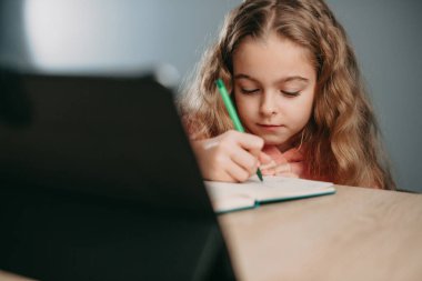 Tablet bilgisayarlı beyaz bir kız evdeki deftere bir şeyler yazıyor. Online eğitim. Çocuk çevrimiçi öğrenim okulu öğreniyor. Karantina, salgın hastalık.