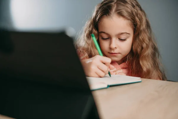 Niña caucásica con la computadora de la tableta de la PC escribiendo a la libreta en casa. Educación en línea. Kid aprender escuela de aprendizaje en línea. Cuarentena, pandemia. —  Fotos de Stock