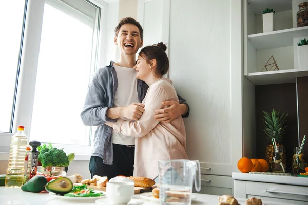 Uomo sorridente abbracciare donna caucasica in piedi in cucina durante il processo di preparazione della colazione. Cucinare, insieme, cucina, relazione. Cibo sano. — Foto Stock