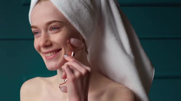 Femme caucasienne souriante en serviette de bain massant le visage avec un rouleau facial en jade pour détendre les muscles. Traitement de rajeunissement. Dermatologie, cosmétologie. Médecine — Video