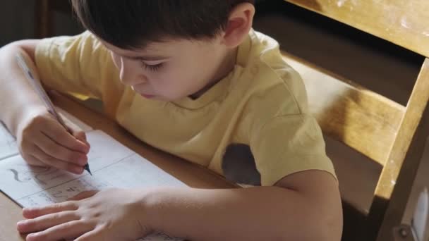 Detailní portrét bělocha sedícího u stolu a cvičícího psaní abecedy do sešitu. Portrét školáka, jak píše dopis. — Stock video