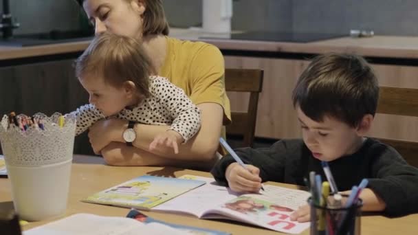 Μητέρα που κρατά το μωρό στην αγκαλιά της ενώ ελέγχει το γιο της, που κάνει τα μαθήματά του, κάθεται στο τραπέζι μαζί της στην κουζίνα. Οικογενειακό Σαββατοκύριακο. Βιβλίο χρωματισμού. — Αρχείο Βίντεο
