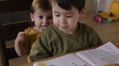 Çocuk, ödevini yaptığı masanın önünde küçük kız kardeşiyle birlikte oturuyor, annesinin eli, çocuğun defterini kapatıyor.