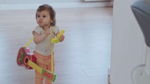 Bebê sentado com guitarra de brinquedo no meio da casa, a mãe levando o instrumento. Menina começando a pisar — Vídeo de Stock