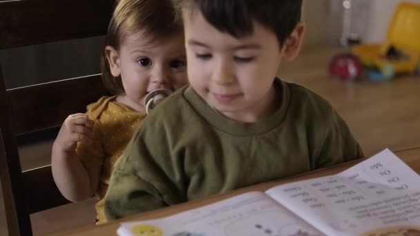 Chlapec sedí se svou mladší sestrou v křesle před stolem, kde si dělal domácí úkoly, a matčina ruka zavírá zápisník chlapce, který — Stock video