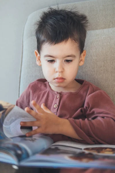 Retrato de cerca de un niño caucásico leyendo un libro sentado en el sofá de su casa. Cuidado familiar. Desarrollo infantil. —  Fotos de Stock