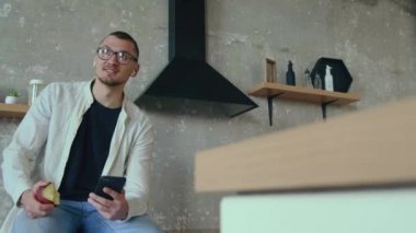 İş adamı cep telefonu kullanarak elma yiyor. İnternetten konuşuyor. Evde tek başına mutfak masasında oturuyor. Çevrimiçi iletişim.