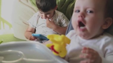 Oturma odasındaki kanepede oturan çocuk akıllı telefon kullanmayı öğrenirken, küçük kız kardeşi de annesi tarafından beslenen yüksek sandalyede oturuyor..