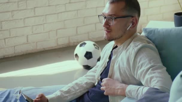 Caucasian man using cellphone and wireless earphones listen to music while sitting on floor in living room. Modern man, great design for any purposes. — Stock Video