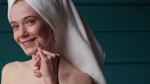 Femme aux taches de rousseur caucasienne avec serviette sur la tête à l'aide d'un rouleau de jade debout dans la salle de bain. Une fille qui rit. Dermatologie, cosmétologie. Traitement de rajeunissement. — Video