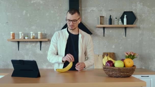 Blanke zakenman neemt deel aan videogesprek op tablet in de keuken tijdens het spelen met een banaan die hij schilt. Moderne levensstijl. — Stockvideo