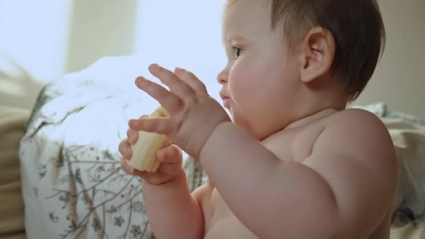 床に座っている間にバナナを食べ裸の赤ん坊の少女のクローズアップ肖像画。ホームコンセプト。ベビーケア. — ストック動画