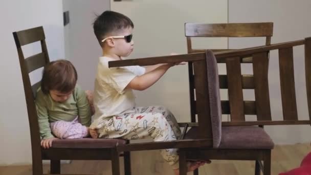 Αγόρι που οδηγάει αυτοσχέδιο αυτοκίνητο στην κουζίνα φορώντας γυαλιά και συνοδευόμενο από την μικρή του αδερφή. Ελεύθερος χρόνος. Η οικογένεια παίζει μαζί. — Αρχείο Βίντεο
