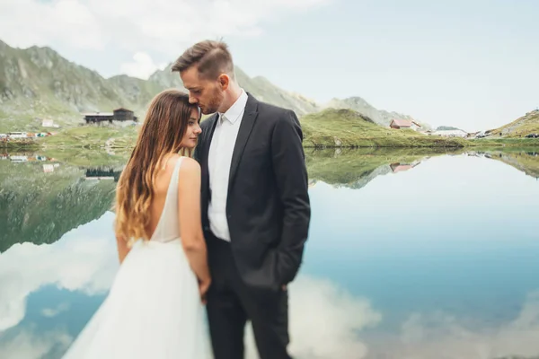 花嫁と新郎湖の背景に立っている。岩の多い山の背景。家庭生活の概念。旅行のコンセプト. — ストック写真