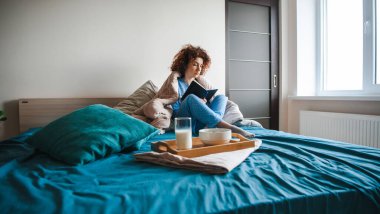 Beyaz saçlı, kıvırcık saçlı bir kadın, sütle mısır gevreği yemeden önce yatakta kitap okuyor. Sağlıklı bir yaşam tarzı. Sağlıklı yemek..