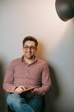 Adam beyaz duvar arkasındayken kitap okuyor. Gözlüklü, gülümseyen beyaz bir öğretmen. Işık arkaplanındaki portre. Duvar arkaplanı