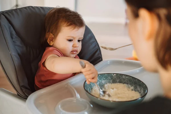 Menina sentada em uma cadeira alta tentando comer mingau sozinho com colher. A mãe a alimentar o bebé. De mãos dadas. — Fotografia de Stock