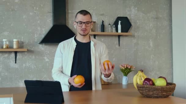Белый мужчина в очках на планшете, в беспроводных наушниках, жонглирует апельсинами. Концепция бизнес образа жизни. Дистанционное образование — стоковое видео