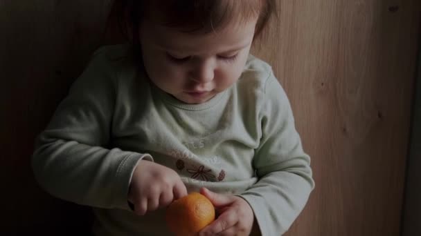 Adorabile bambino che cerca di sbucciare un mandarino seduto a casa sul pavimento. Mani in alto. Cura del bambino. Assistenza alla famiglia. Ritratto ravvicinato. — Video Stock
