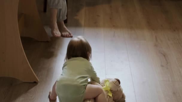 Vista posteriore di una bambina seduta in cucina che sale sul peluche orsacchiotto dal pavimento, cavalcando. Bella ragazza. — Video Stock
