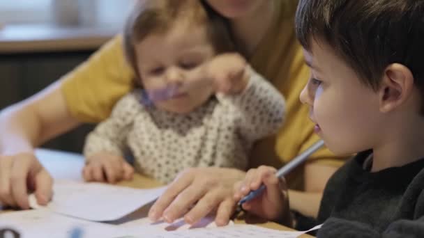 Niño haciendo la tarea con la ayuda de su padre cariñoso. Mamá ayudando a su hijo con la tarea mientras sostiene a su hija en brazos. Estudiar, hacer los deberes. — Vídeo de stock