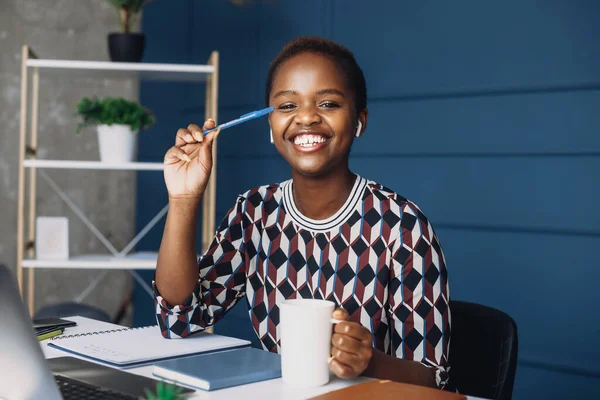 Retrato de un hermoso trabajador afro-americano sonriente sentado en el escritorio, mirando a la cámara, usando una computadora portátil. Hermosa mujer africana. Retrato de mujer de negocios — Foto de Stock