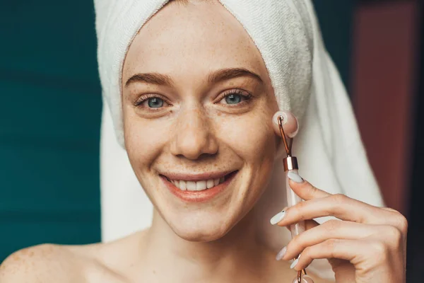 Ginger vrouw met handdoek op hoofd masseren gezicht met behulp van jade roller. Medische behandeling. — Stockfoto