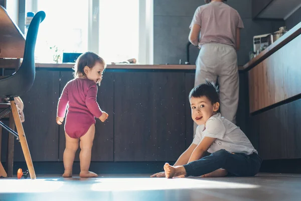 Due bambini piccoli che giocano seduti sul pavimento di legno in cucina a casa godendosi il tempo libero divertendosi. Famiglia felice. — Foto Stock