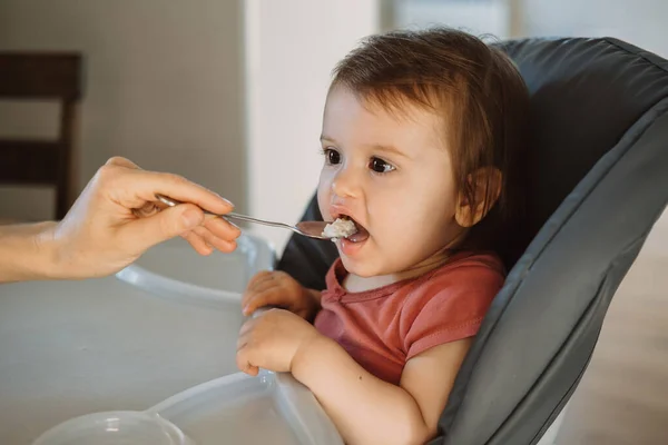 Мать кормит ребенка ложкой, девочка сидит дома и ест пищу на стуле. Счастливая семья. Улыбающийся счастливый ребенок. — стоковое фото
